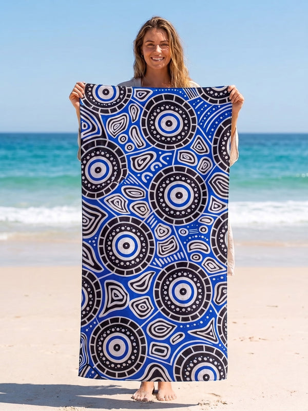 A woman standing on a sandy beach holds up a large rectangular towel or blanket featuring a bold blue, black, white, and gray circular dot-art pattern. The ocean and blue sky are visible in the background.