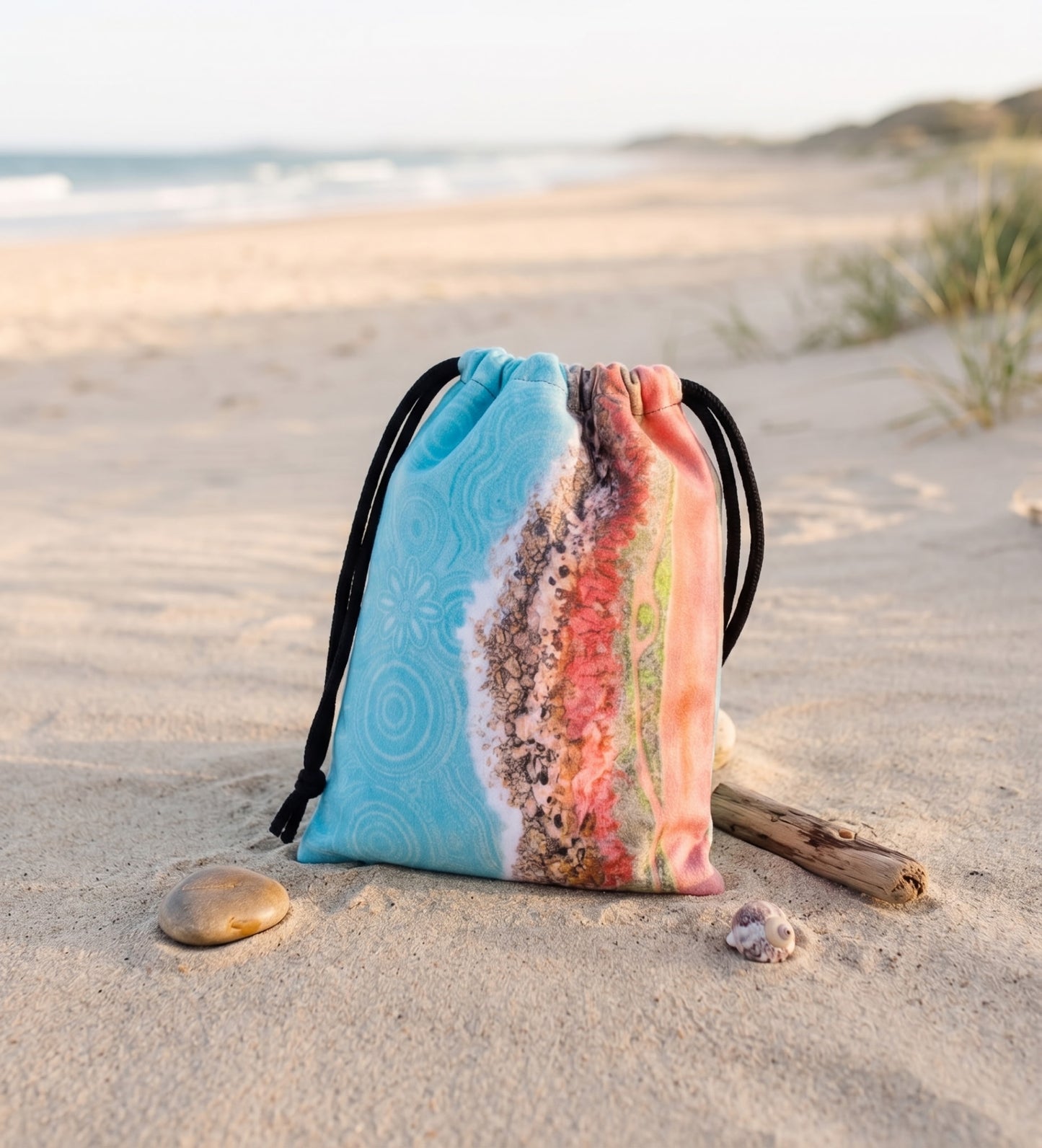 A drawstring beach bag with a colorful printed design featuring light blue, pink, and brown tones, placed on a sandy beach next to a piece of driftwood.