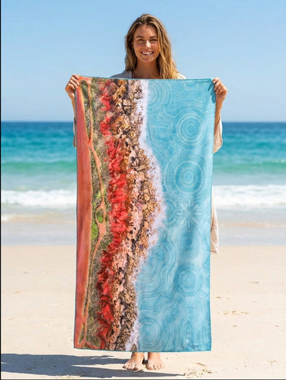 A woman standing on a sandy beach holds up a travel towel featuring a split design: one side shows a red, orange, and brown abstract floral or textured pattern, and the other side is light blue with circular wave-like patterns.